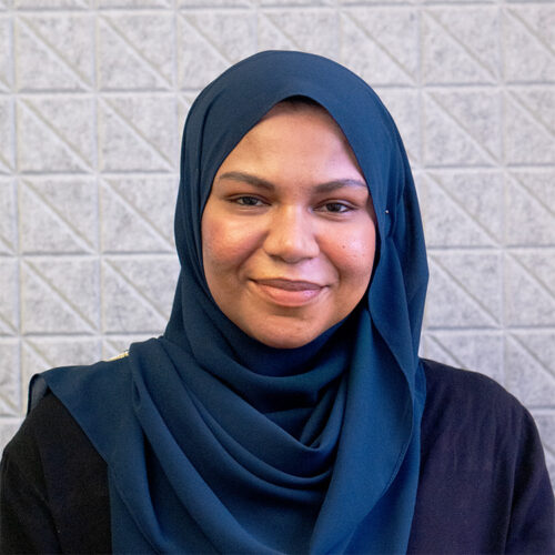 Smiling woman wearing a dark blue hijab against a light gray tiled wall